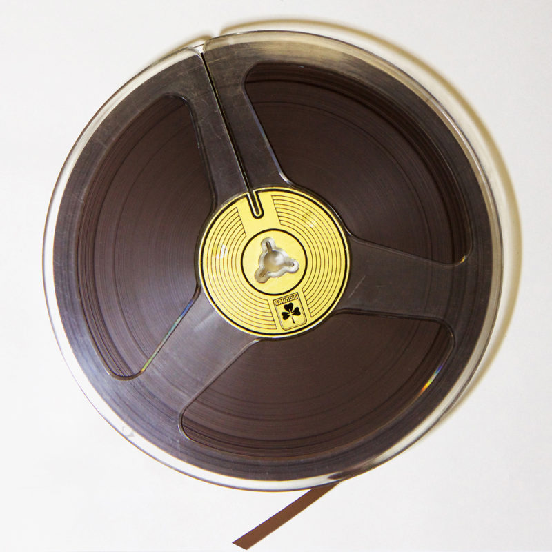 Clear plastic 7 inch reel containing quarter inch brown magnetic audio tape. In the centre of the reel is gold Grundig trefoil label.