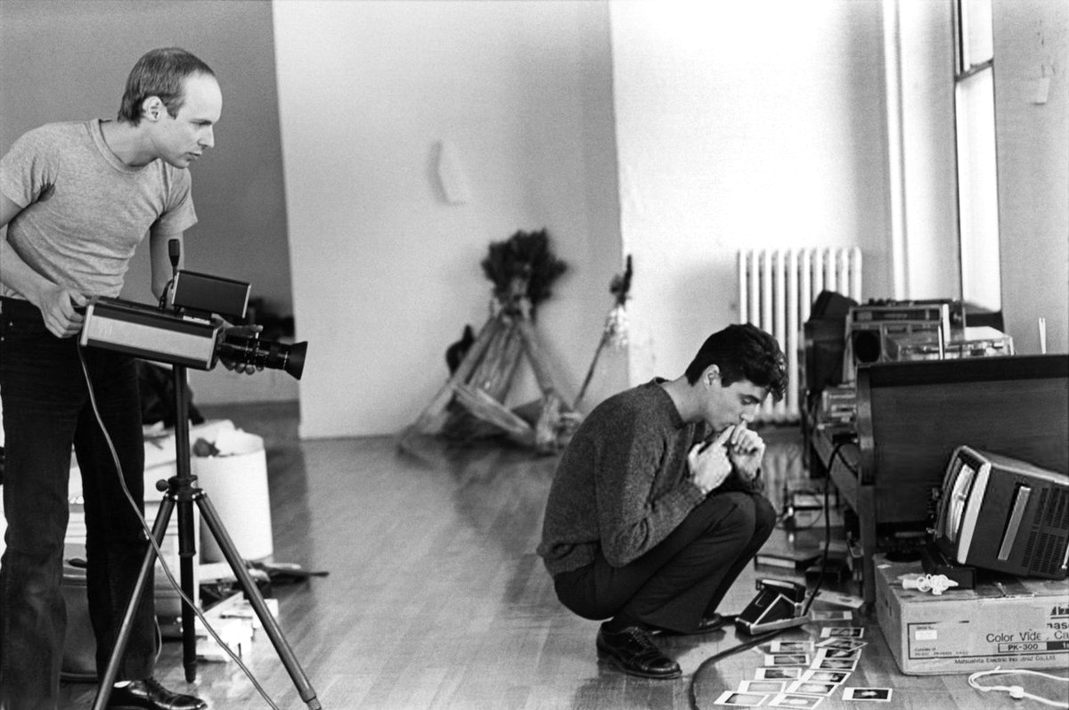 late 1970s black and white of Brian Eno and David Byrne with large video camera sideways on tripod. Eno watching monitor, Byrne inspecting polaroids